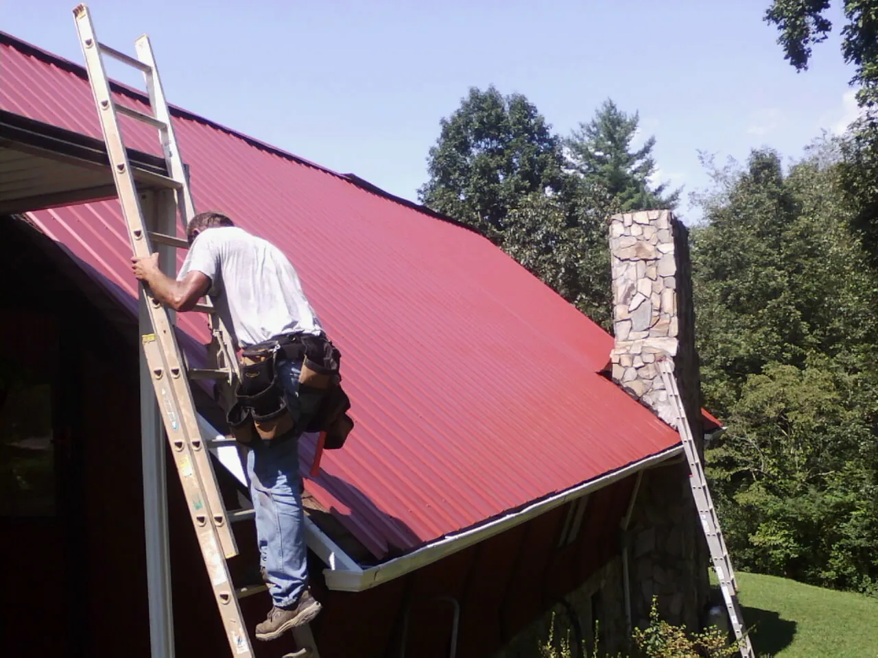 Roofer on a ladder beside a red metal roof and stone chimney — Soffit & Fascia Repair by Roofing Brothers in Andrew